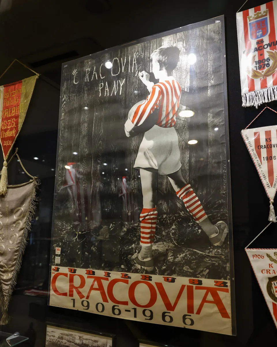 Jubileuszowy plakat „Cracovia 1906–1966” z rysunkiem piłkarza w klubowych barwach, eksponowany obok proporczyków w muzealnej gablocie.