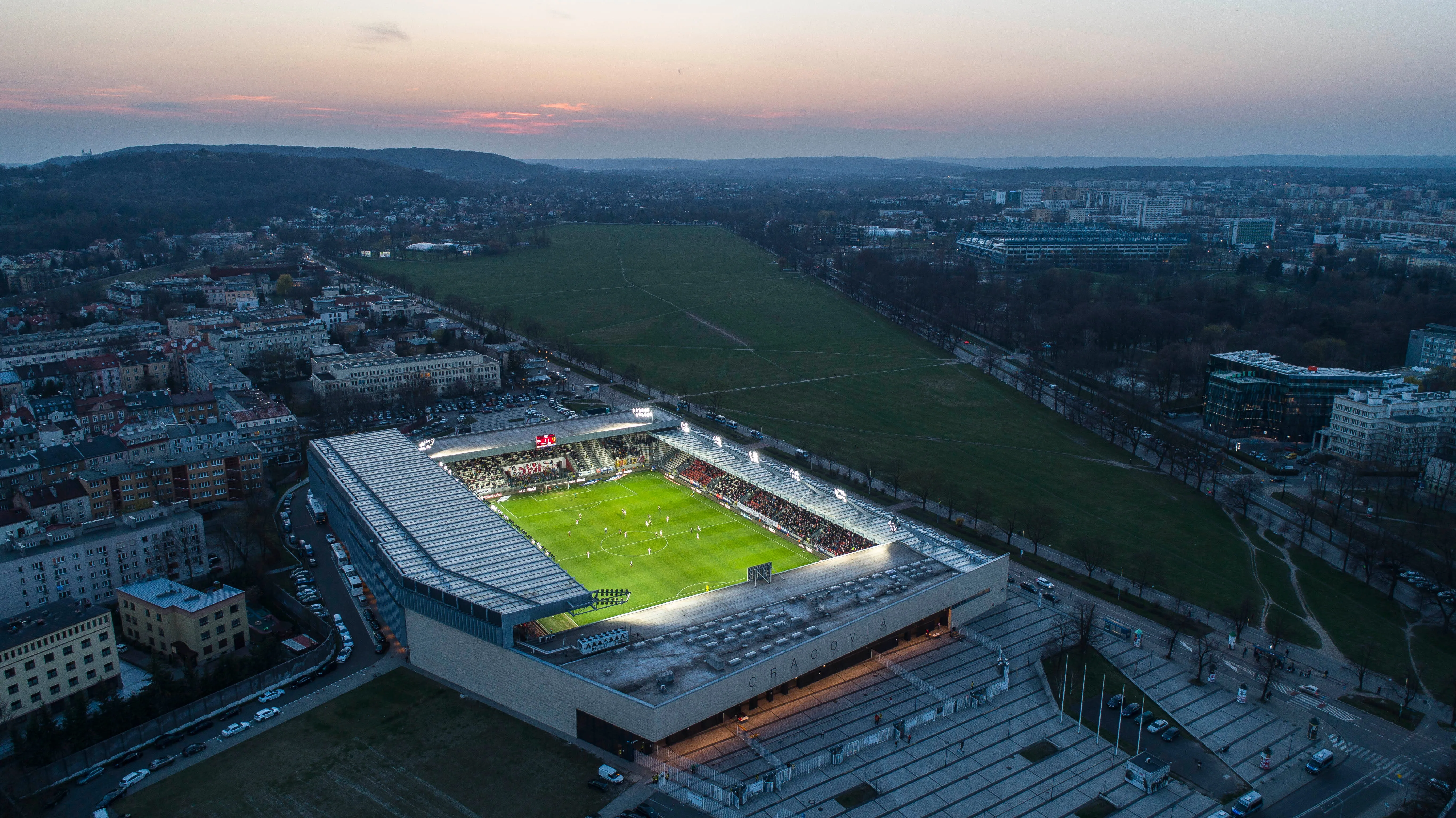 Widok z lotu ptaka na stadion Cracovii i otaczającą go zabudowę Krakowa o zachodzie słońca.